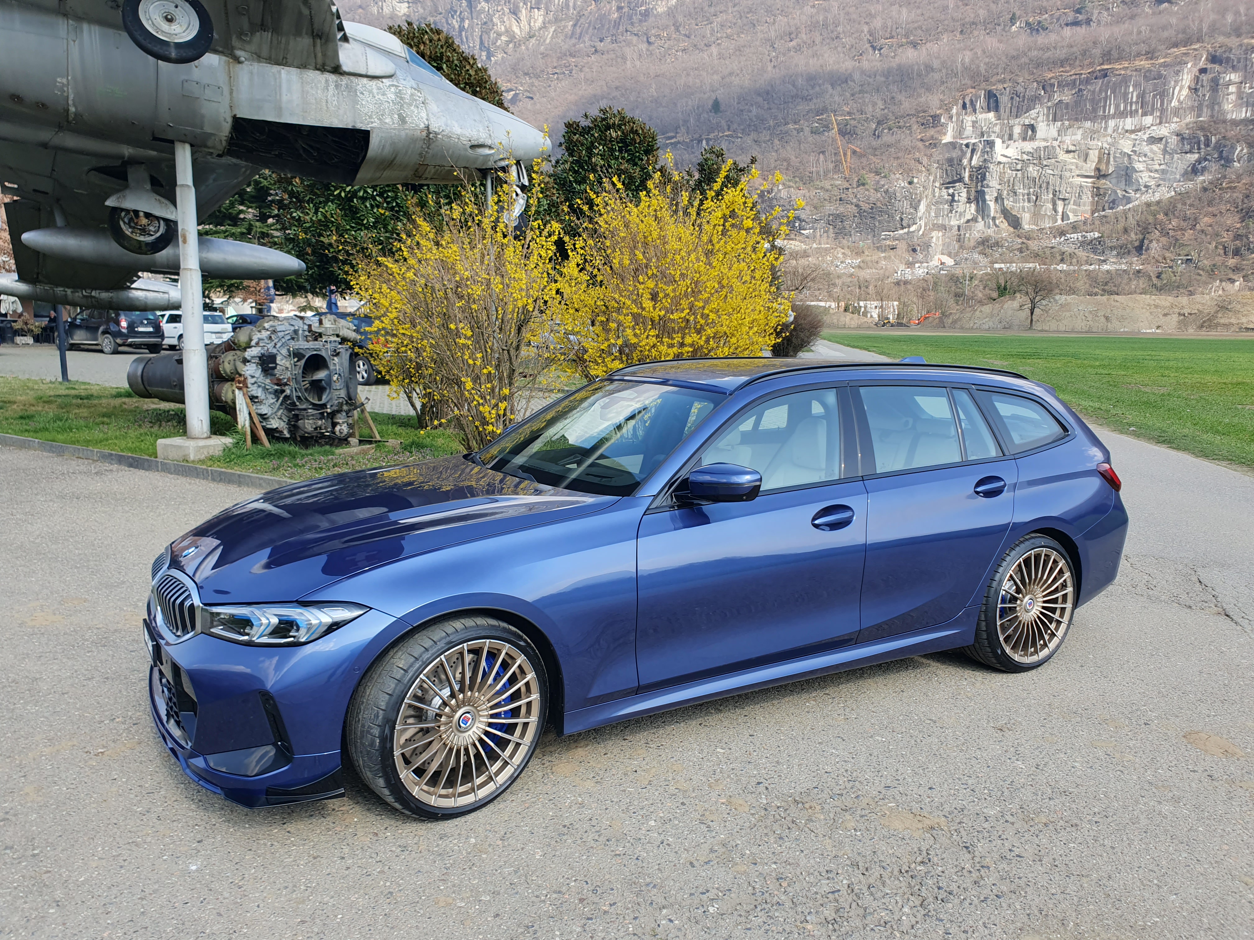 Μπλε μεταλλικό BMW Alpina Touring με χάλκινες ζάντες, σταθμευμένο σε εξωτερικό χώρο μπροστά από έκθεμα στρατιωτικού αεροσκάφους.